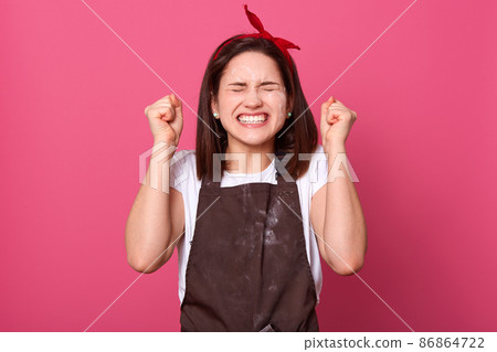 Housewife female wearing brown apron and white casual t shirt, standing isolated over rose wall background. Happy expressive woman doing winner gesture, clenching her fist, celebrates succses. 86864722