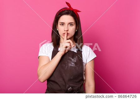 Close up portrait of young charming girl with finger on her lips, makes silence gesture, female wears red hairband, white casual t shirt, brown apron, looks directly at camera, has serious expression. 86864726