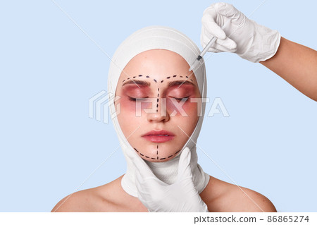 Attractive young woman with closed eyes against blue studio wall. Female in bandage and with surgical lines being injected by doctor. Lady dislikes her wrinkles on forehead. Plastic surgery concept. 86865274