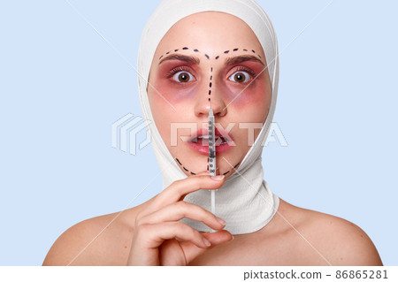 Portrait of woman in clinic with bandage on head. Lady holds syringe. Lady with opened mouth looks scared isolated over blue background. Girl afraids of injection. Beauty and cosmetic surgery concept. 86865281