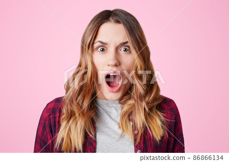 Shocked stupefied female student, opens mouth widely, being surprised to know that tomorrow is final exam, wears checkered shirt, isolated over pink background. Negative human emotions concept Shocked stupefied female student, opens mouth widely, being surprised to know that tomorrow is final exam, wears checkered shirt, isolated over pink background. Negative human emotions concept 86866134