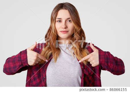 Horizontal shot of attractive woman with wavy hair, points at blank space of her clothes, advertises new outfit, wears checkered shirt, isolated over white background. People and design concept Horizontal shot of attractive woman with wavy hair, points at blank space of her clothes, advertises new outfit, wears checkered shirt, isolated over white background. People and design concept 86866155