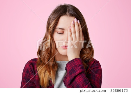 Isolated shot of pretty frustrated overworked female covers face with hand, keeps eyes shut, being sleepy, feels bored as spends time alone, poses against pink background. Tiredness concept. Isolated shot of pretty frustrated overworked female covers face with hand, keeps eyes shut, being sleepy, feels bored as spends time alone, poses against pink background. Tiredness concept. 86866173