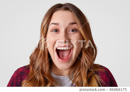 Close up portrait of happy young European woman opens mouth widely, has long wavy hair, being overjoyed, isolated over white background. Stylish lady expresses good emotions. Happiness concept Close up portrait of happy young European woman opens mouth widely, has long wavy hair, being overjoyed, isolated over white background. Stylish lady expresses good emotions. Happiness concept 86866175