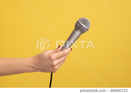Profile side view closeup of human hand holding silver microphone for singing or reporting. Indoor studio shot isolated on yellow background. 86866725