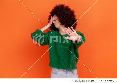 Depressed woman with Afro hairstyle wearing green casual style sweater covering eyes with palm and showing stop ban gesture with palm. Indoor studio shot isolated on orange background. Depressed woman with Afro hairstyle wearing green casual style sweater covering eyes with palm and showing stop ban gesture with palm. Indoor studio shot isolated on orange background. 86866769