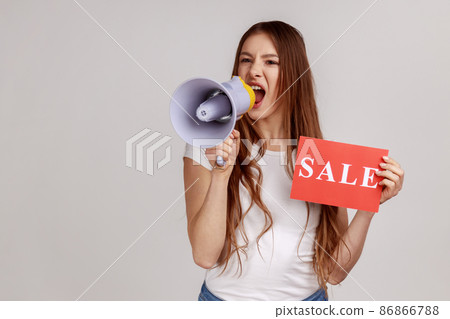 Portrait of excited attractive young adult woman holding sale card and megaphone, announcing discounts in shopping mall, wearing white T-shirt. Indoor studio shot isolated on gray background. 86866788