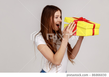 Excited woman with dark hair unboxing present box, looking inside with surprised facial expression, birthday gift, wearing white T-shirt. Indoor studio shot isolated on gray background. Excited woman with dark hair unboxing present box, looking inside with surprised facial expression, birthday gift, wearing white T-shirt. Indoor studio shot isolated on gray background. 86866830