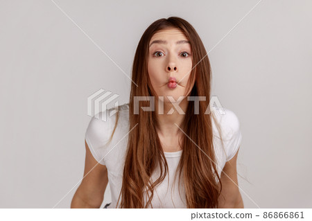 Surprised funny woman with dark hair making fish face grimace with pout lips and looking with confused comical expression, wearing white T-shirt. Indoor studio shot isolated on gray background. Surprised funny woman with dark hair making fish face grimace with pout lips and looking with confused comical expression, wearing white T-shirt. Indoor studio shot isolated on gray background. 86866861