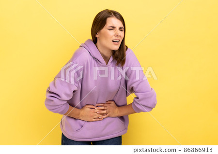 Portrait of unhealthy distressed woman clutching her belly, grimacing from acute abdominal pain, gastritis or constipation, wearing purple hoodie. Indoor studio shot isolated on yellow background. Portrait of unhealthy distressed woman clutching her belly, grimacing from acute abdominal pain, gastritis or constipation, wearing purple hoodie. Indoor studio shot isolated on yellow background. 86866913
