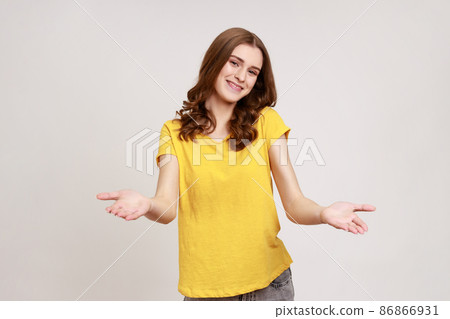 Please, take for free. Friendly pretty young woman in yellow T-shirt welcoming with wide open arms and smiling kindly, looking smiling at camera. Indoor studio shot isolated on gray background. 86866931