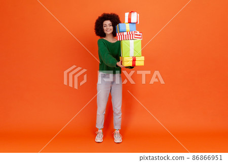 Full length portrait of woman with Afro hairstyle wearing green casual style sweater holding present boxes, looking at camera with toothy smile. Indoor studio shot isolated on orange background. Full length portrait of woman with Afro hairstyle wearing green casual style sweater holding present boxes, looking at camera with toothy smile. Indoor studio shot isolated on orange background. 86866951