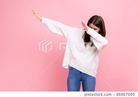 Woman with dark hair showing popular internet meme pose, celebrating success victory, dabbing trends, wearing white casual style sweater. Indoor studio shot isolated on pink background. 86867009