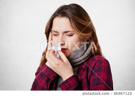 Sick female blows her nose in tissue, looks ill and exhausted, has terrible headache, running nose, caught cold during cold winter weather. Beautiful young woman has flu and thore throat Sick female blows her nose in tissue, looks ill and exhausted, has terrible headache, running nose, caught cold during cold winter weather. Beautiful young woman has flu and thore throat 86867128