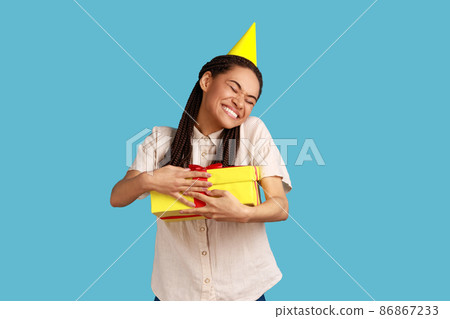 Happy woman with black dreadlocks in party cone holding embracing yellow wrapped present box, being satisfied to get gift, wearing white shirt. Indoor studio shot isolated on blue background. Happy woman with black dreadlocks in party cone holding embracing yellow wrapped present box, being satisfied to get gift, wearing white shirt. Indoor studio shot isolated on blue background. 86867233