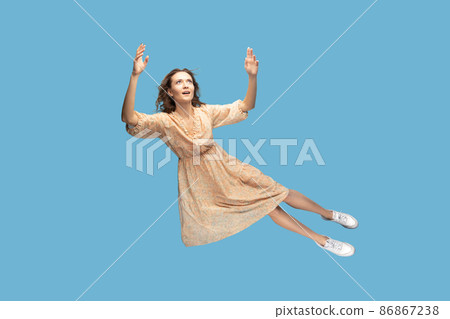 Hovering in air. Full length beautiful pensive girl in yellow dress levitating flying in mid-air, looking up with dreamy relaxed expression. indoor studio shot isolated on blue background Hovering in air. Full length beautiful pensive girl in yellow dress levitating flying in mid-air, looking up with dreamy relaxed expression. indoor studio shot isolated on blue background 86867238