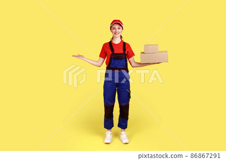 Full length portrait of optimistic delivery woman standing with cardboard packages and presenting copy space on palm, wearing overalls and red cap. Indoor studio shot isolated on yellow background. Full length portrait of optimistic delivery woman standing with cardboard packages and presenting copy space on palm, wearing overalls and red cap. Indoor studio shot isolated on yellow background. 86867291