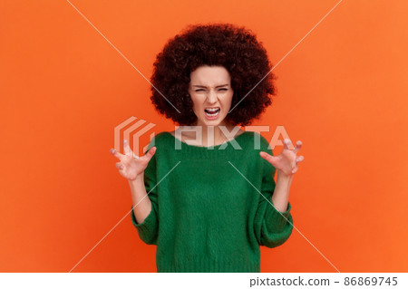 Portrait of angry woman with Afro hairstyle wearing green casual style sweater expressing aggressive emotions, raised arms, screaming. Indoor studio shot isolated on orange background. Portrait of angry woman with Afro hairstyle wearing green casual style sweater expressing aggressive emotions, raised arms, screaming. Indoor studio shot isolated on orange background. 86869745