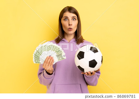 Portrait of woman with open mouth holding soccer ball and fan of euro, amazed of football championship, betting, wearing purple hoodie. Indoor studio shot isolated on yellow background. 86869781