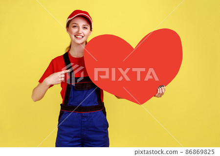 Smiling happy worker woman pointing at big red heart with finger, looking at camera, expressing positive emotions, wearing overalls and red cap. Indoor studio shot isolated on yellow background. Smiling happy worker woman pointing at big red heart with finger, looking at camera, expressing positive emotions, wearing overalls and red cap. Indoor studio shot isolated on yellow background. 86869825