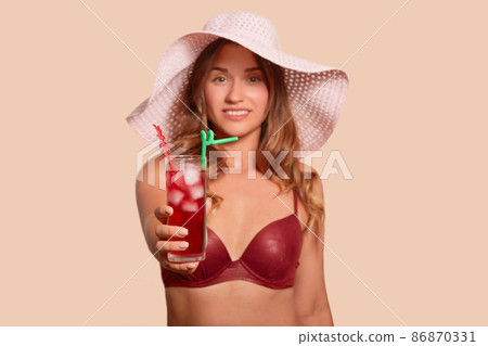 Image of smiling charismatic lady raising one arm, holding glass of red drink with ice, being in good mood, looking directly at camera, being on vacation, wearing light hat and swimming suit. Image of smiling charismatic lady raising one arm, holding glass of red drink with ice, being in good mood, looking directly at camera, being on vacation, wearing light hat and swimming suit. 86870331