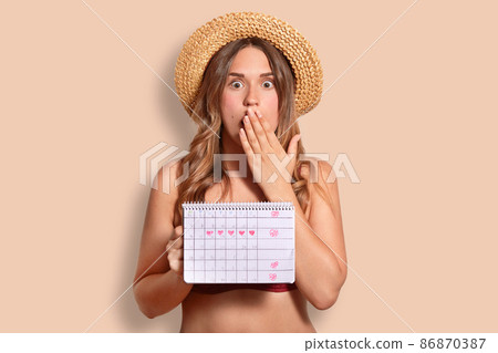 Shocked terrified female covers mouth with palm, shcoked to have periods, holds period calendar, wears bikini and straw hat, isolated over studio background. People, summer rest and women concept 86870387