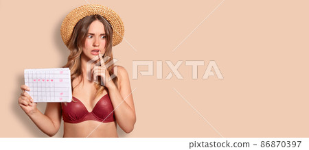 Indoor shot of thoughtful embarrassed female wears summer hat and swimsuit, holds period calendar, tries to remember date of periods, poses against beige background with copy space for your text 86870397