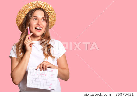 Horizontal shot of positive dreamy female holds marker and periods calendar, being in high spirit, isolated over pink background with copy space for your promotional content or advertisement 86870431