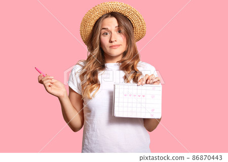 Hesitant young lady carries menstruation period calendar, has clueless expression, wears headgear and white casual t shirt, isolated over pink wall, hesitates about something. Gynecological concept 86870443