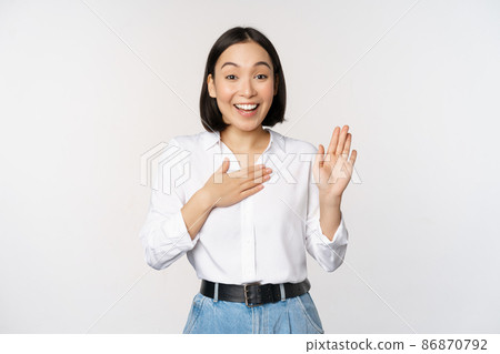 Image of cute young female office worker, asian girl student raising hand up and put palm on chest, name herself, introduce, making promise, standing over white background 86870792