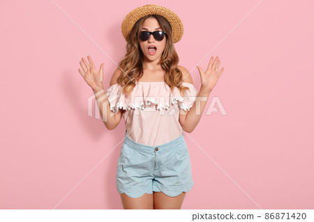 Image of young woman wearing pink blouse, blue short, sunglasses and summer straw hat standing with open mouth in shock, keeps hands up, has astonished facial expression. Human emotions concept. Image of young woman wearing pink blouse, blue short, sunglasses and summer straw hat standing with open mouth in shock, keeps hands up, has astonished facial expression. Human emotions concept. 86871420