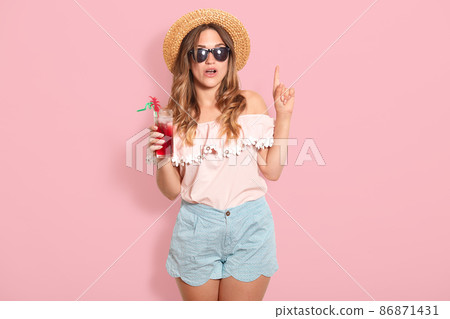 Attractive young woman in summer outfit isolated on pink background. Happy girl in sunglasses posing with straw hat and cocktail, spending hot summer days on beach, points fore finger up, has new idea Attractive young woman in summer outfit isolated on pink background. Happy girl in sunglasses posing with straw hat and cocktail, spending hot summer days on beach, points fore finger up, has new idea 86871431