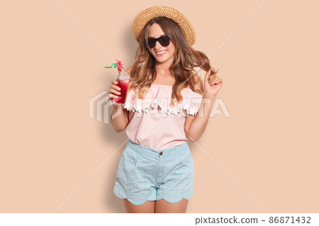Beautiful cheerful female rests in resort country, holds summer fresh cocktail, wears trendy sunglasses, blouse and shorts, poses against studio wall. People, lifestyle, recreation and rest concept Beautiful cheerful female rests in resort country, holds summer fresh cocktail, wears trendy sunglasses, blouse and shorts, poses against studio wall. People, lifestyle, recreation and rest concept 86871432