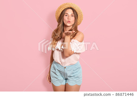 Close up portrait of beautiful woman wearing straw hat, blouse, short, stands smiling and blowing air kiss directly at camera isolated over pink background in studio, posing with bared shoulders. Close up portrait of beautiful woman wearing straw hat, blouse, short, stands smiling and blowing air kiss directly at camera isolated over pink background in studio, posing with bared shoulders. 86871446
