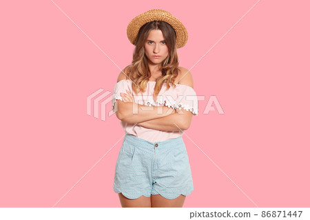 Attractive upset female with frowned face standing over pink background in studio, having folded arms, looking directly at camera, being disappointed, wearing blouse, blue shorts and straw hat. Attractive upset female with frowned face standing over pink background in studio, having folded arms, looking directly at camera, being disappointed, wearing blouse, blue shorts and straw hat. 86871447