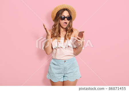 Beautiful astonished woman wearing blouse with bared shoulders, short, straw hat and sunglasses, holding smart phone in hand and posing with open mouth, recives shocking messages from friend. 86871455