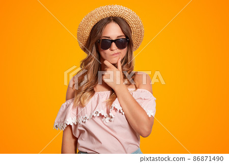 Thoughtful pensive beautiful female holds chin, has hesitant expression, wears shades and summer hat, dressed in shoulder sleeved blouse, poses against orange background. People and rest concept Thoughtful pensive beautiful female holds chin, has hesitant expression, wears shades and summer hat, dressed in shoulder sleeved blouse, poses against orange background. People and rest concept 86871490