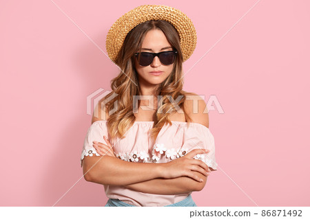 Close up portrait of young fair haired woman wearing pink shirt, straw hat and sunglasses, holding her hands folded and looking seriously at camera, looks offended, isolated over pink background. Close up portrait of young fair haired woman wearing pink shirt, straw hat and sunglasses, holding her hands folded and looking seriously at camera, looks offended, isolated over pink background. 86871492