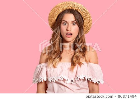 Portrait of shocked Caucasian woman wears straw hat and fashionable blouse, shows bare shoulders, isolated over pink background, surprised to have spoiled vacation. Omg and reaction concept. Portrait of shocked Caucasian woman wears straw hat and fashionable blouse, shows bare shoulders, isolated over pink background, surprised to have spoiled vacation. Omg and reaction concept. 86871494