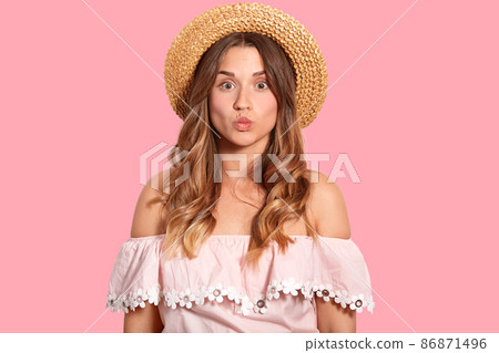 Beautiful European woman with long wavy hair, pouts lips, makes grimace, wears straw hat and summer blouse, has healthy skin, poses against pink background, ready for holidays. Horizontal shot Beautiful European woman with long wavy hair, pouts lips, makes grimace, wears straw hat and summer blouse, has healthy skin, poses against pink background, ready for holidays. Horizontal shot 86871496