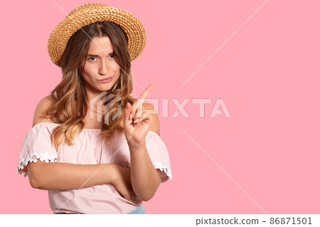 Horizontal shot of serious curious woman with long hair, dressed in fashionable straw hat and blouse, points with fore finger at blank space for your advertising content or promotion. Look there! 86871501