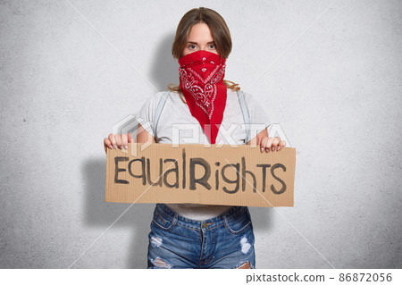 Horizontal shot of beautiful young female belong to feminist move, wears red bandana on face, holds plate with inscription Equal rights, demonstrates womens power, stands indoor alone, isolated 86872056