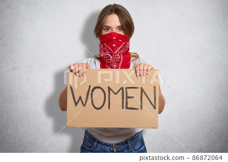 Horizontal shot of beautiful feminist holds plate with inscription Women, belong to special move, has face covered with red badana, isolated over grey concrete wall. Feminism and women concept 86872064
