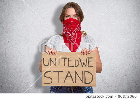 Womens right concept. Attractive serious young woman holds plate, wears casual clothes and red stylish bandana on face, belongs to feminist move, defenses females, isolated over white background Womens right concept. Attractive serious young woman holds plate, wears casual clothes and red stylish bandana on face, belongs to feminist move, defenses females, isolated over white background 86872066