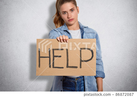Cropped image of adorable female dressed in denim clothes, holds plate with inscription Help, defenses females rights, poses against grey concrete wall. People, power, feminism and women concept Cropped image of adorable female dressed in denim clothes, holds plate with inscription Help, defenses females rights, poses against grey concrete wall. People, power, feminism and women concept 86872087