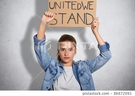 Horizontal shot of serious young woman shows fist, dressed in denim jacket, holds plate above head, being self confident in womens power, isolated over white concrete wall. Feminism concept. 86872091
