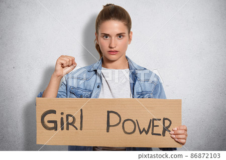 Lovely confident young woman with serious expression, shows fists, demonstares her power, belongs to feminists, wears denim jacket, isolated over white wall. Feminism, rights and equality concept 86872103