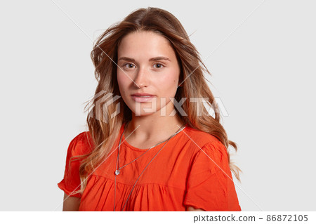 Headshot of gorgeous lovely young female with healthy skin, wavy luxurious hair, dressed in orange sun dress, looks directly at camera, isolated over white background. People and beauty concept 86872105