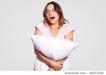 Photo of tired attractive female model yawns, opens mouth widely, dressed in casual clothes, carries pillow, needs to have good rest, isolated over white background. Woman hugs cushion indoor 86872449