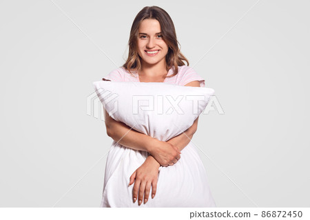 Image of positive Caucasian lady with toothy smile hugs pillow, wears pajamas, poses in studio against white background. Happy young woman in good mood after having pleasant dreams at night. Image of positive Caucasian lady with toothy smile hugs pillow, wears pajamas, poses in studio against white background. Happy young woman in good mood after having pleasant dreams at night. 86872450